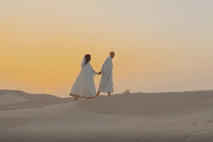 Un couple en tenues blanches traditionnelles marche main dans la main sur une dune de sable au coucher du soleil